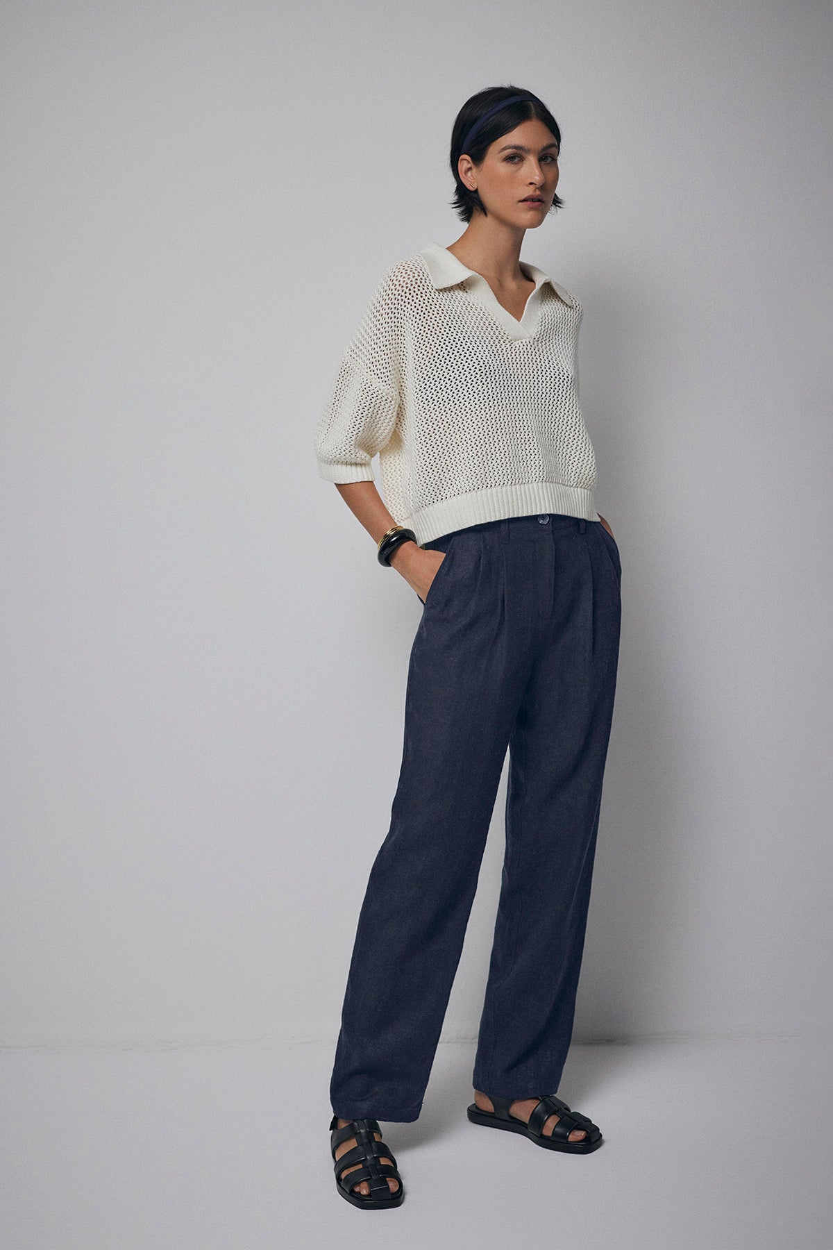   A person stands against a plain white background, exuding casual charm in a Velvet by Jenny Graham YORK SWEATER POLO, dark blue trousers, black sandals, and two black bracelets. 