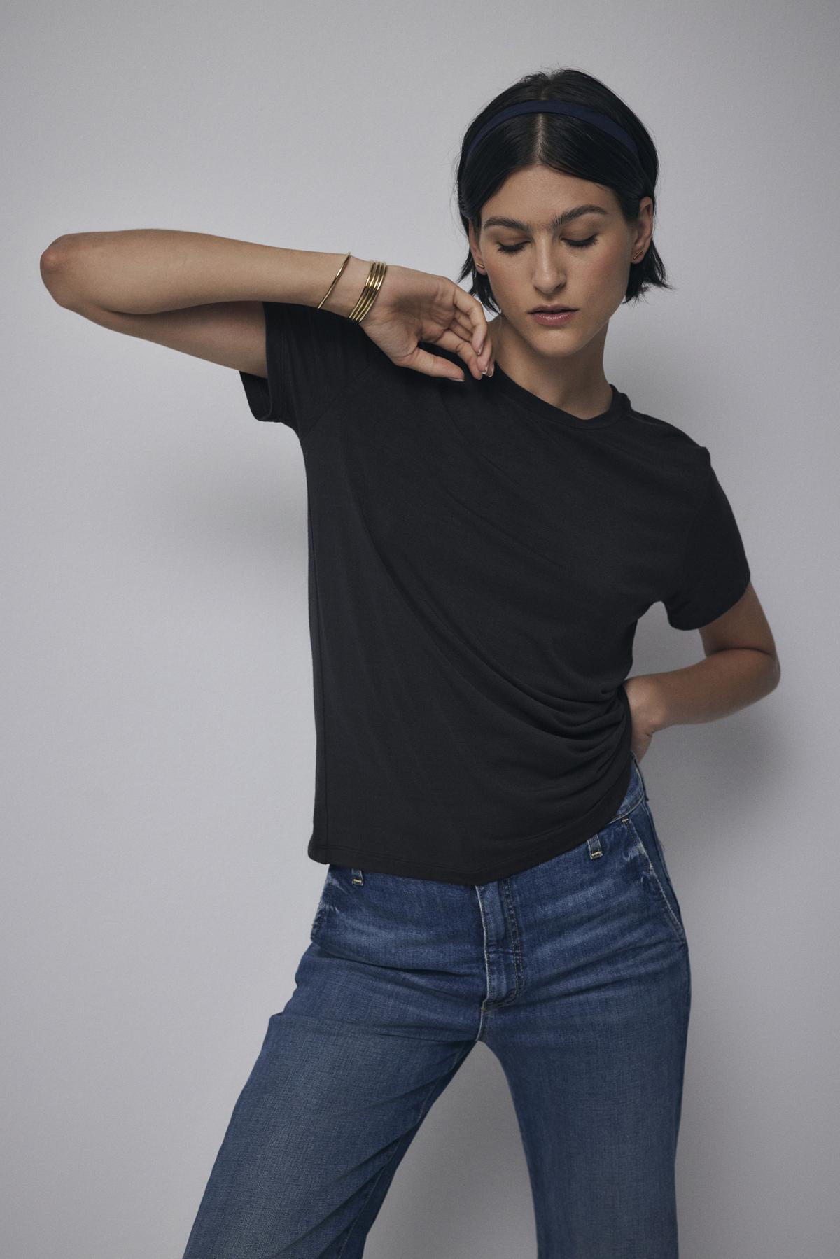   A person with short dark hair wearing a classic black Velvet by Jenny Graham SOLANA TEE and tailored blue jeans poses against a plain background. 