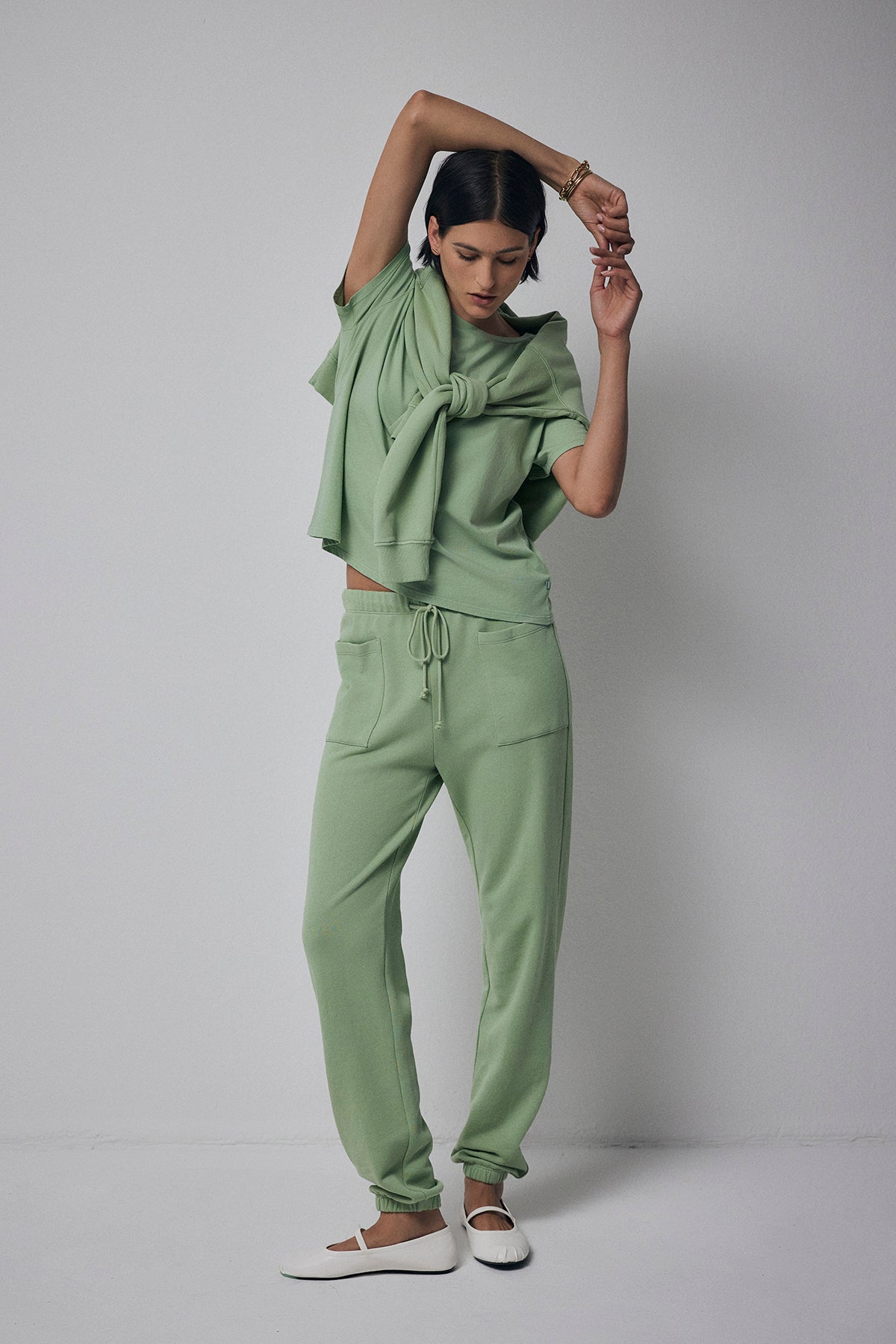   A person models the WESTLAKE SWEATPANT by Velvet by Jenny Graham, showcasing a coordinated sage green outfit with a tied top and relaxed silhouette drawstring pants. The elastic drawstring waist of the sweatpants ensures comfort. They complement their look with white shoes and strike a pose against a plain background, one hand raised above their head while looking down. 