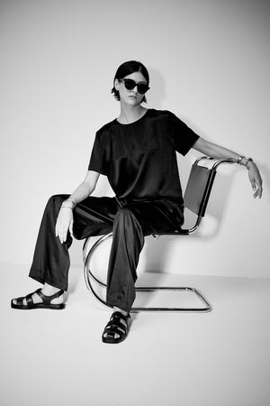 A person in black Velvet by Jenny Graham Manhattan Pant and sunglasses sitting confidently on a modern chair against a white background.