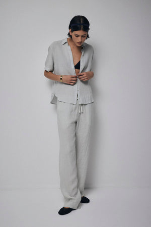 Woman in a PICO LINEN PANT from Velvet by Jenny Graham, with relaxed fit pants and black top, standing and looking down at her phone, against a plain white backdrop.