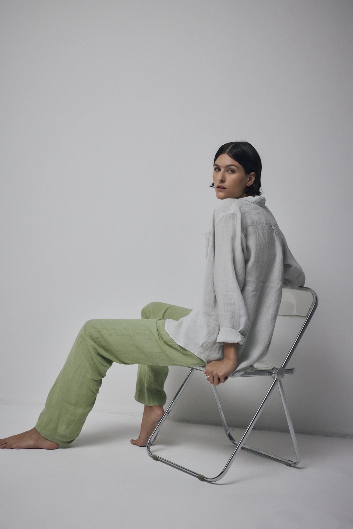   A woman sits on a metal chair against a plain background, wearing a white blouse and Velvet by Jenny Graham's PICO LINEN PANT in green, looking away thoughtfully. 