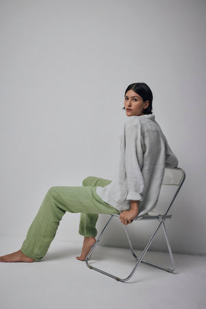 Woman sitting on a metal chair in a neutral-colored room, wearing a loose gray top and relaxed fit green Velvet by Jenny Graham PICO PANT.