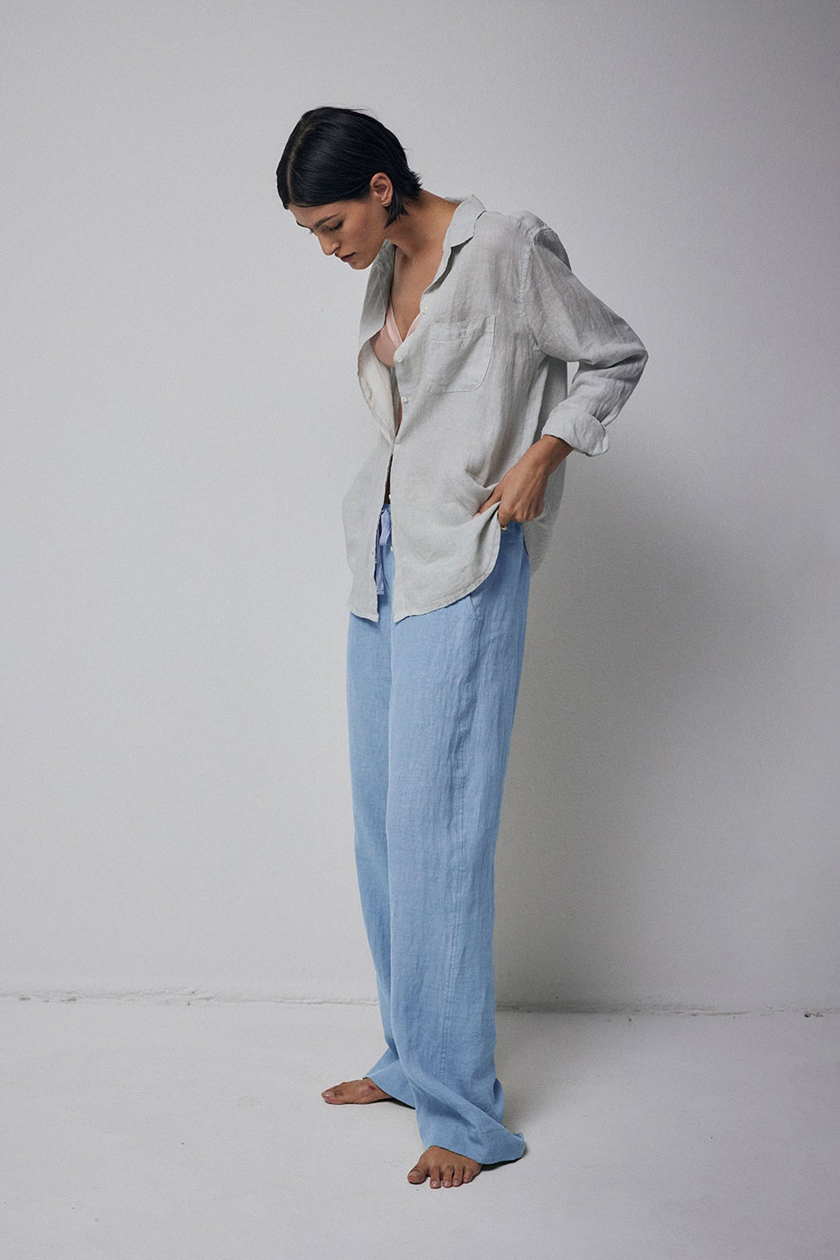 A person standing barefoot in a studio, wearing light blue Velvet by Jenny Graham Pico Pants and a loosely fitted grey shirt.-36212496859329