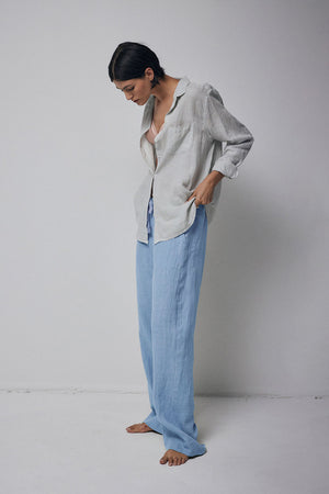 A person standing barefoot in a studio, wearing light blue Velvet by Jenny Graham Pico Pants and a loosely fitted grey shirt.