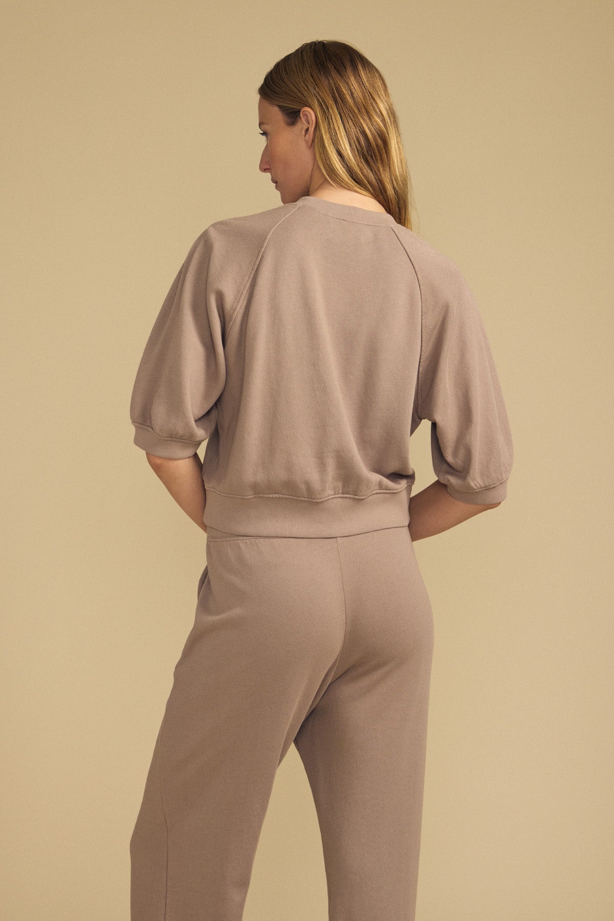 A person stands with their back to the camera, wearing the BONNIE SWEATSHIRT by Velvet by Graham & Spencer in taupe, paired with matching pants against a neutral beige background.-41814383624385
