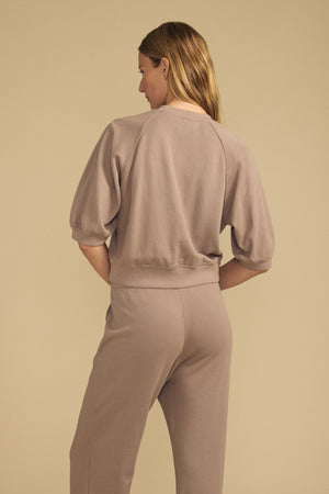 A person stands with their back to the camera, wearing the BONNIE SWEATSHIRT by Velvet by Graham & Spencer in taupe, paired with matching pants against a neutral beige background.