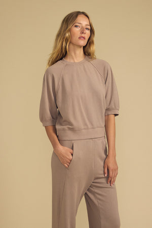 A woman wearing the BONNIE SWEATSHIRT by Velvet by Graham & Spencer, styled in taupe with matching pants, stands with one hand in her pocket against a beige background.