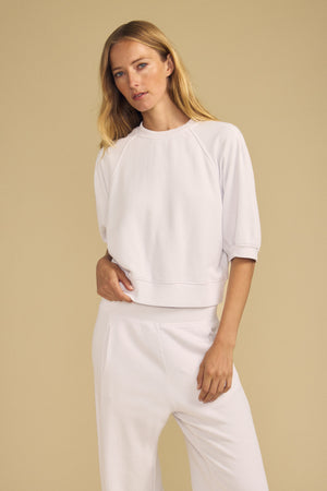 A woman with long blonde hair wears the BONNIE SWEATSHIRT by Velvet by Graham & Spencer—a white top with elbow-length sleeves—paired with matching white pants, standing against a beige background.