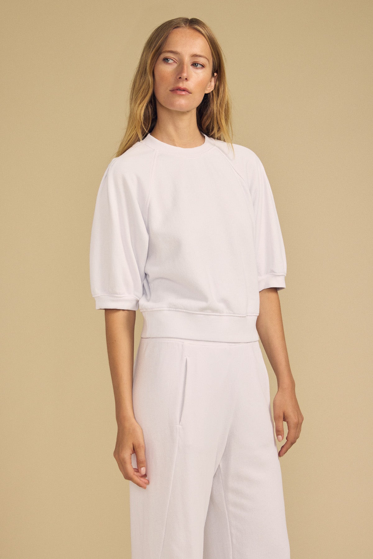 A woman stands against a beige background wearing the BONNIE SWEATSHIRT by Velvet by Graham & Spencer with matching white pants, looking slightly to the side.-41814383427777