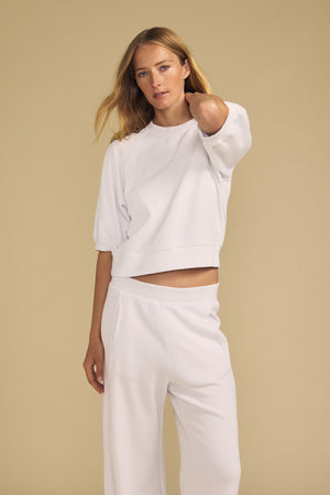 A woman with long blonde hair wears the BONNIE SWEATSHIRT and matching wide-leg pants by Velvet by Graham & Spencer, standing against a plain beige background.