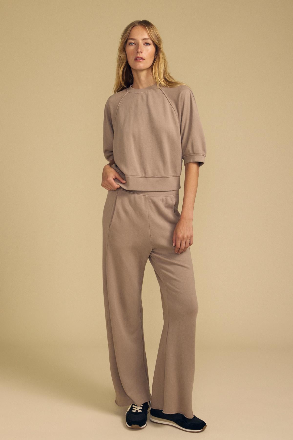 A woman stands against a beige background wearing the Velvet by Graham & Spencer QHILLA SWEATPANT matching set in taupe, featuring half sleeves and wide-leg sweatpants, paired with black sneakers.-41834186670273