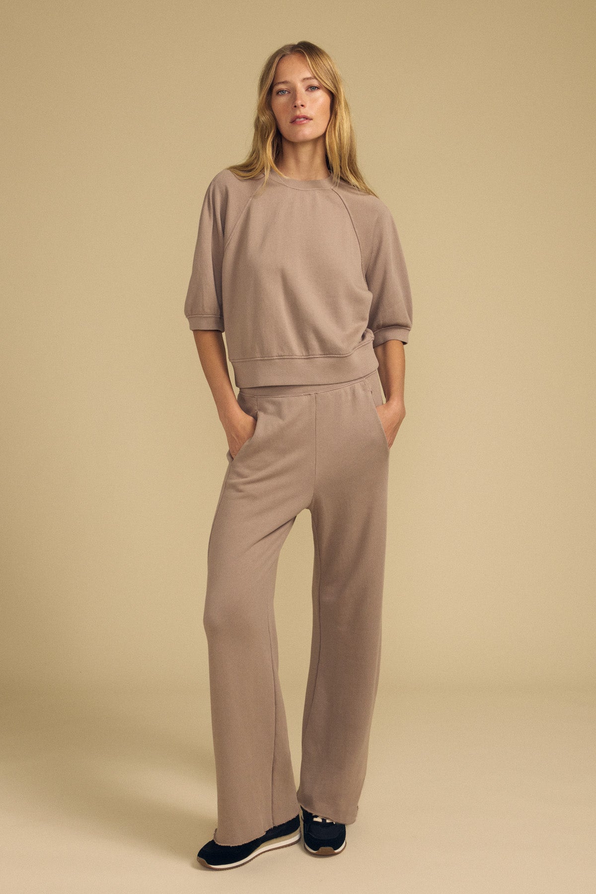 A woman stands against a beige background wearing the Velvet by Graham & Spencer QHILLA Sweatpant— a loose-fitting, wide-leg taupe cotton fleece set paired with black sneakers, her hands tucked in her pockets.