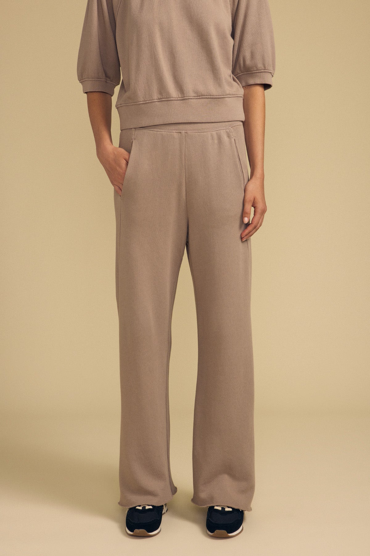 A person is wearing the QHILLA SWEATPANT by Velvet by Graham & Spencer, a taupe cotton fleece wide leg sweatpant paired with a matching sweatshirt, hands in pockets and black sneakers, standing against a plain beige background.