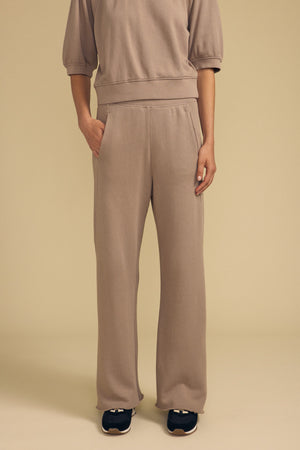 A person is wearing the QHILLA SWEATPANT by Velvet by Graham & Spencer, a taupe cotton fleece wide leg sweatpant paired with a matching sweatshirt, hands in pockets and black sneakers, standing against a plain beige background.