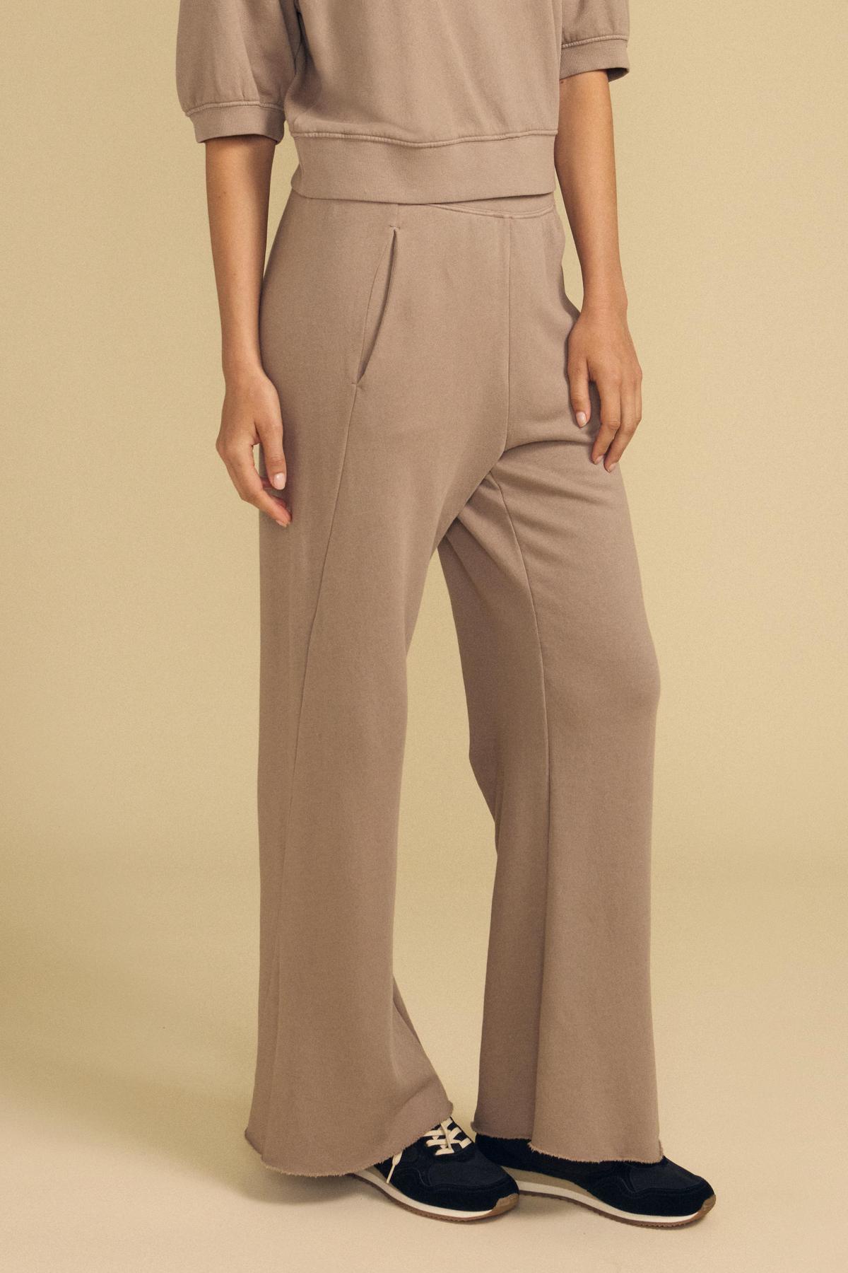 A person is seen standing against a beige background, wearing the Velvet by Graham & Spencer QHILLA SWEATPANT in taupe paired with black sneakers; the upper part of their body is partially out of frame.