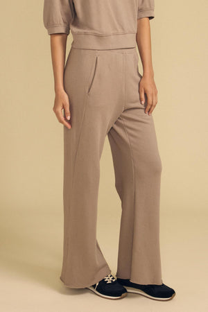 A person is seen standing against a beige background, wearing the Velvet by Graham & Spencer QHILLA SWEATPANT in taupe paired with black sneakers; the upper part of their body is partially out of frame.