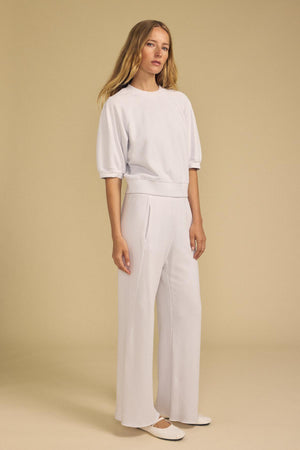 A woman stands against a beige background wearing a white short-sleeve top, white shoes, and the QHILLA SWEATPANT by Velvet by Graham & Spencer.