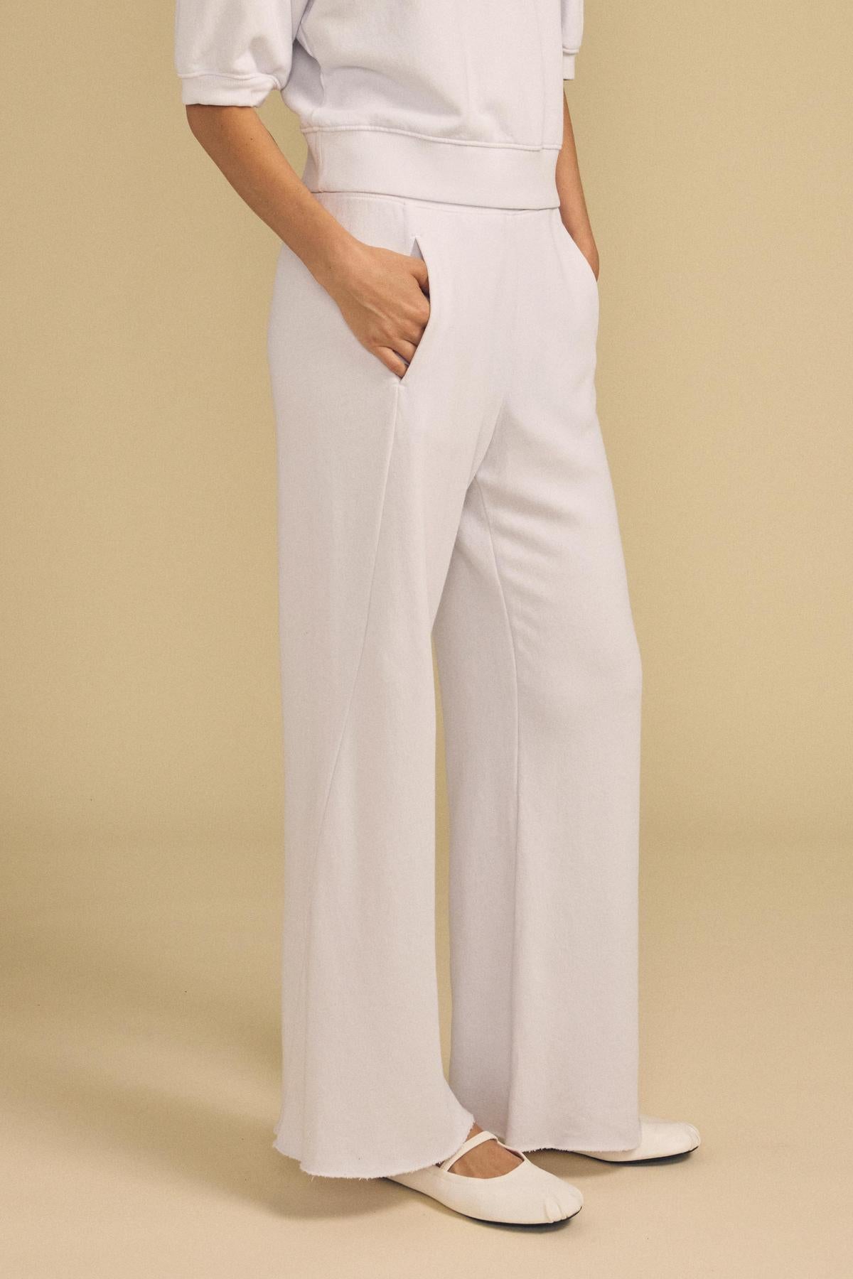 Wearing the QHILLA SWEATPANT by Velvet by Graham & Spencer in white with hands in pockets, paired with white ballet flats, standing against a beige background.