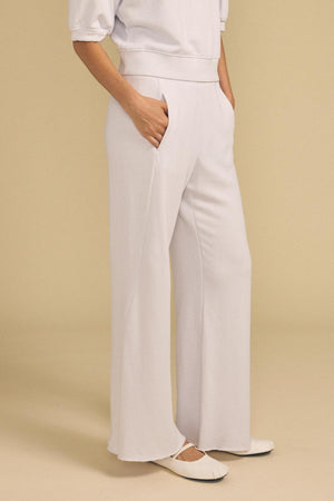 Wearing the QHILLA SWEATPANT by Velvet by Graham & Spencer in white with hands in pockets, paired with white ballet flats, standing against a beige background.
