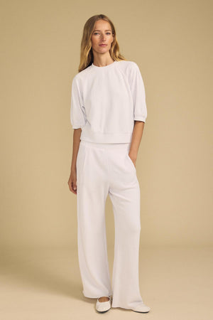 A woman is standing against a beige background, wearing the Velvet by Graham & Spencer QHILLA SWEATPANT—a white, loose-fitting matching set featuring wide-leg sweatpants and a half-sleeve top.