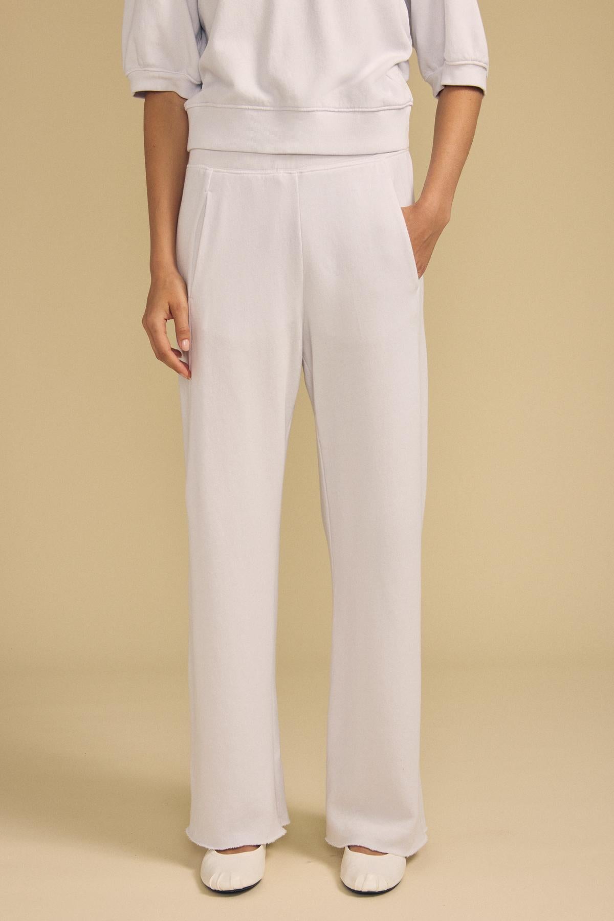 A person stands against a beige background, wearing the Velvet by Graham & Spencer QHILLA SWEATPANT in a relaxed fit, paired with a white sweatshirt and white ballet flats. Only the lower torso and legs are visible.-41940416233665