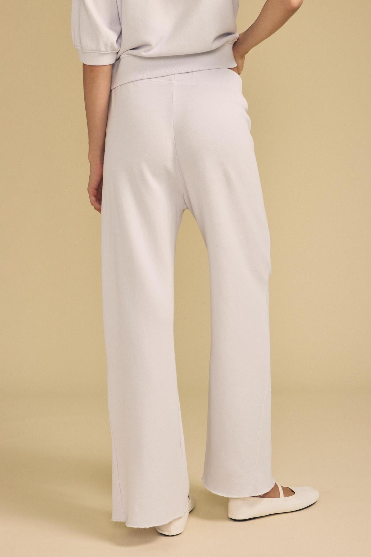Wearing the Velvet by Graham & Spencer QHILLA SWEATPANT in a loose, light-colored wide leg silhouette, paired with white flats, the person stands on a beige background with their back facing the camera.