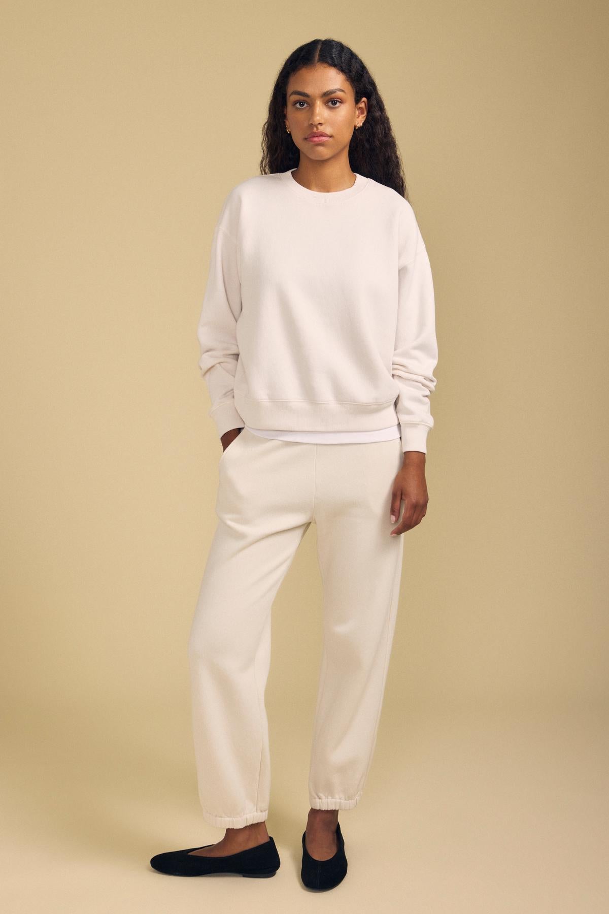 A person stands facing forward, wearing the YOKO SWEATSHIRT by Velvet by Graham & Spencer in a relaxed fit cream-colored cotton fleece, paired with matching sweatpants and black slip-on shoes, against a plain beige background.-41965236420801