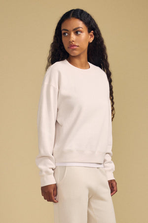 A woman with long curly hair stands against a beige background, looking to her left. She wears the cream-colored YOKO SWEATSHIRT by Velvet by Graham & Spencer and matching pants. The relaxed fit and cropped design of the sweatshirt lend effortless style to her look.