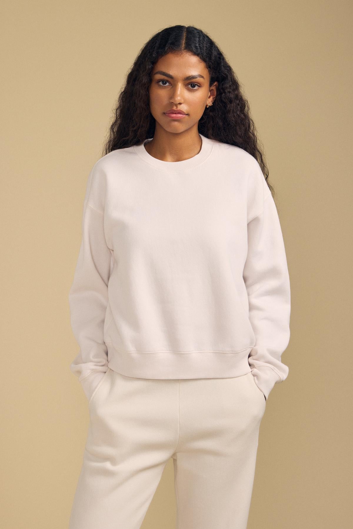 A woman with long curly hair, hands in her pockets, stands against a plain beige background wearing the relaxed fit YOKO SWEATSHIRT by Velvet by Graham & Spencer in light beige cotton fleece with matching pants.