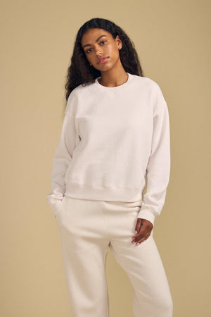 A person wearing the Velvet by Graham & Spencer YOKO SWEATSHIRT in a relaxed off-white cotton fleece, paired with matching pants, stands against a plain beige background with one hand in their pocket.