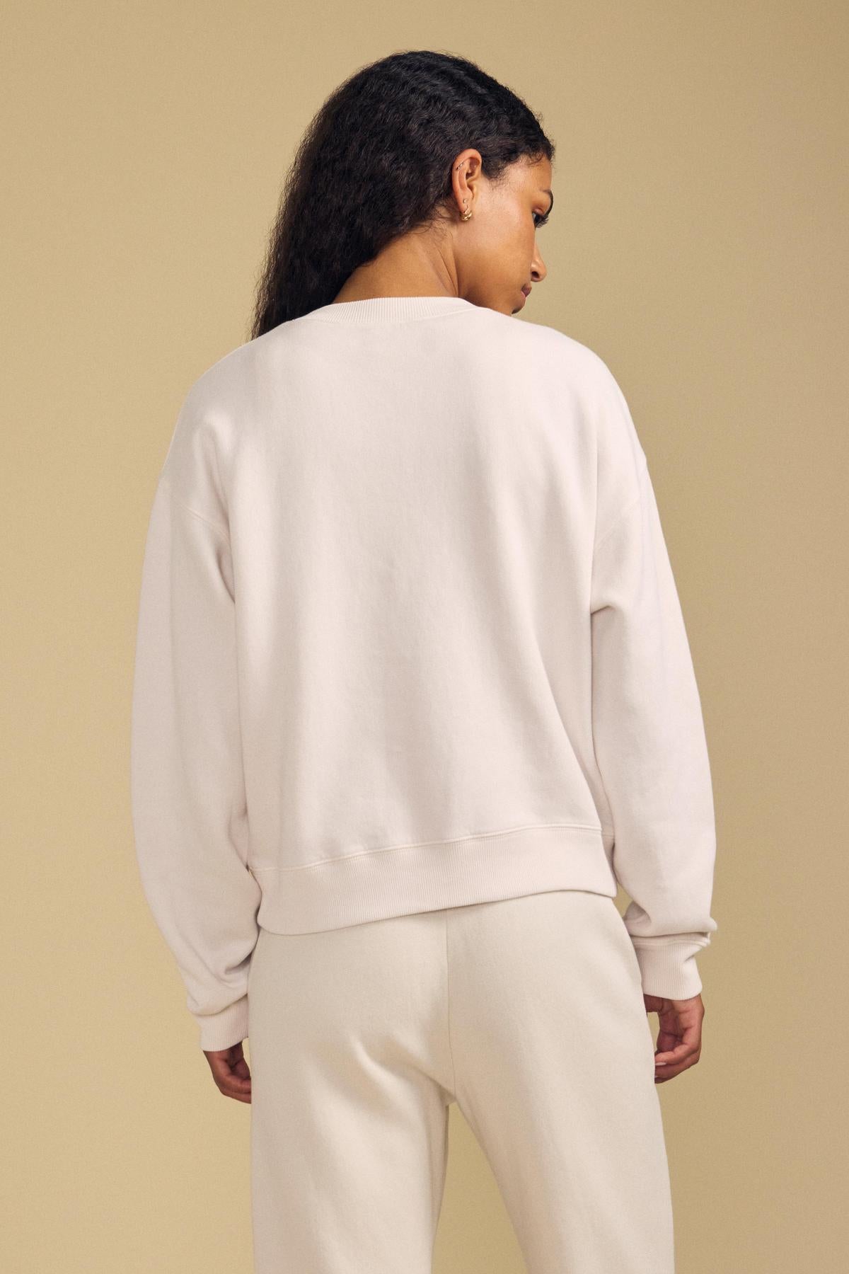 A person with long, curly hair is seen from behind against a beige background, wearing the YOKO SWEATSHIRT by Velvet by Graham & Spencer along with relaxed fit cotton fleece pants.