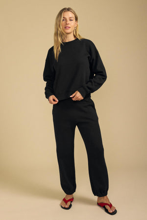 A person stands against a beige background wearing a black sweatshirt, relaxed fit black ZARA SWEATPANT by Velvet by Graham & Spencer in cotton fleece, and red-and-black sandals.