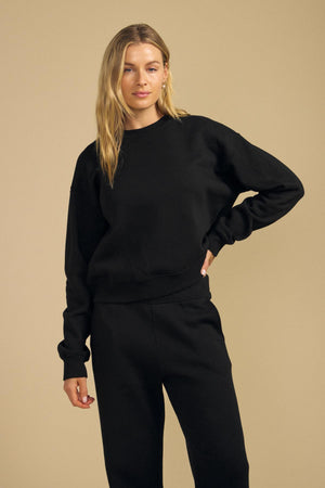 A woman with long blond hair wears the Velvet by Graham & Spencer YOKO SWEATSHIRT and matching black pants, both crafted from soft cotton fleece, while standing against a plain beige background.