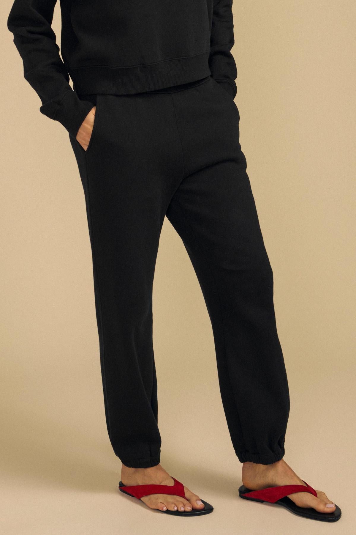 Wearing the ZARA SWEATPANT from Velvet by Graham & Spencer in a relaxed black jogger style, paired with red flip-flops, this person stands against a beige background with hands in their pockets.