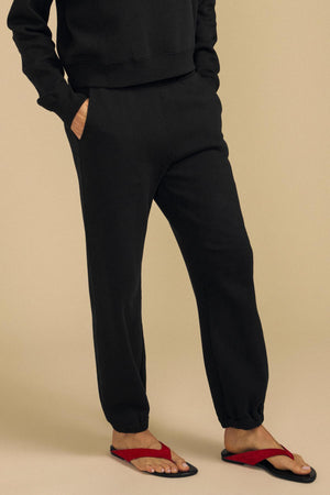Wearing the ZARA SWEATPANT from Velvet by Graham & Spencer in a relaxed black jogger style, paired with red flip-flops, this person stands against a beige background with hands in their pockets.
