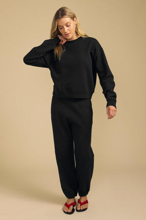 A person wearing the YOKO SWEATSHIRT by Velvet by Graham & Spencer in black, paired with matching black sweatpants, stands on a beige background looking down with one hand touching their face. Crafted from soft cotton fleece, the outfit features a relaxed silhouette. They complete the look with red sandals.
