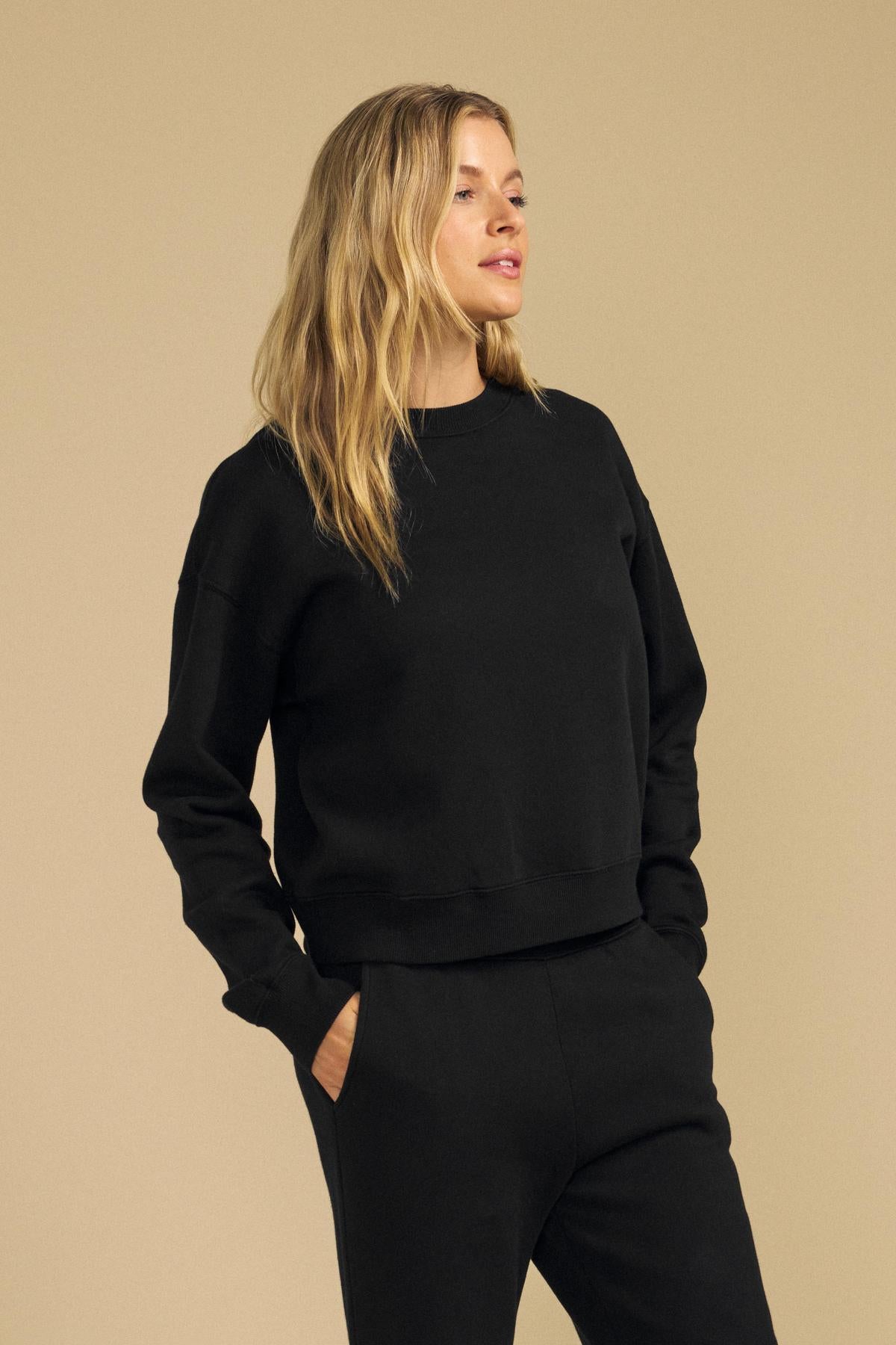 A woman with long blonde hair wears the YOKO Sweatshirt and matching black pants from Velvet by Graham & Spencer, their relaxed silhouette and soft cotton fleece adding effortless comfort as she stands with one hand in her pocket against a beige background.-41980700197057