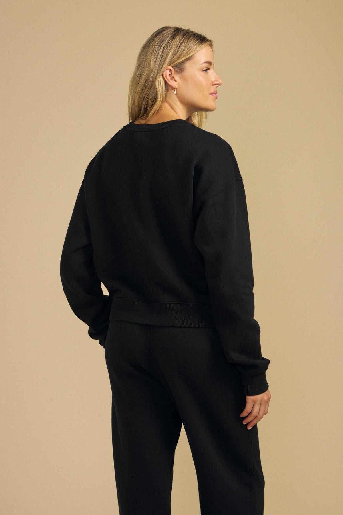 A woman with blonde hair stands sideways against a plain beige background, wearing the Velvet by Graham & Spencer YOKO SWEATSHIRT in cotton fleece, styled in a relaxed silhouette, paired with black pants. She gazes slightly to the left.
