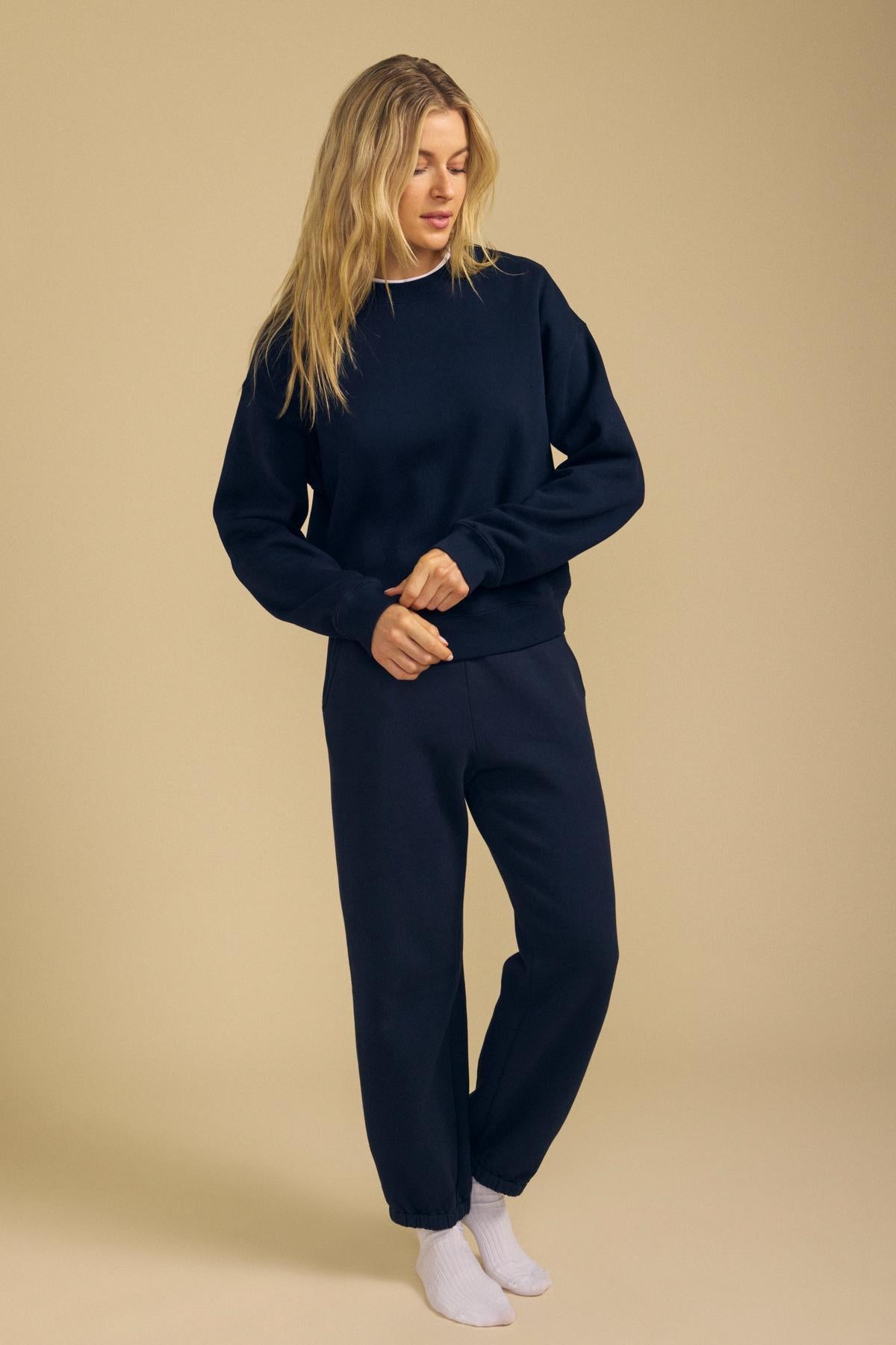 Standing against a beige background, the model wears a navy blue relaxed fit sweatshirt paired with matching ZARA SWEATPANT by Velvet by Graham & Spencer in soft cotton fleece, complemented by white socks. She looks down and holds one sleeve.-42187958386881
