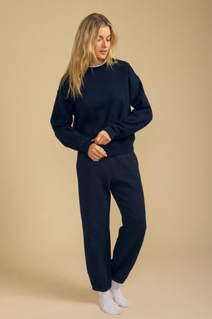 Standing against a beige background, the model wears a navy blue relaxed fit sweatshirt paired with matching ZARA SWEATPANT by Velvet by Graham & Spencer in soft cotton fleece, complemented by white socks. She looks down and holds one sleeve.