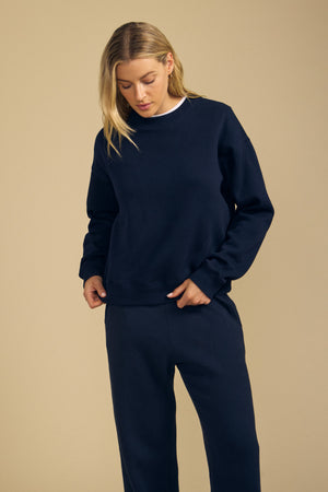 A person wearing a navy blue YOKO SWEATSHIRT by Velvet by Graham & Spencer and matching pants stands against a plain beige background, looking down with one hand near their waist. The cotton fleece fabric and relaxed silhouette create an effortlessly comfortable look.