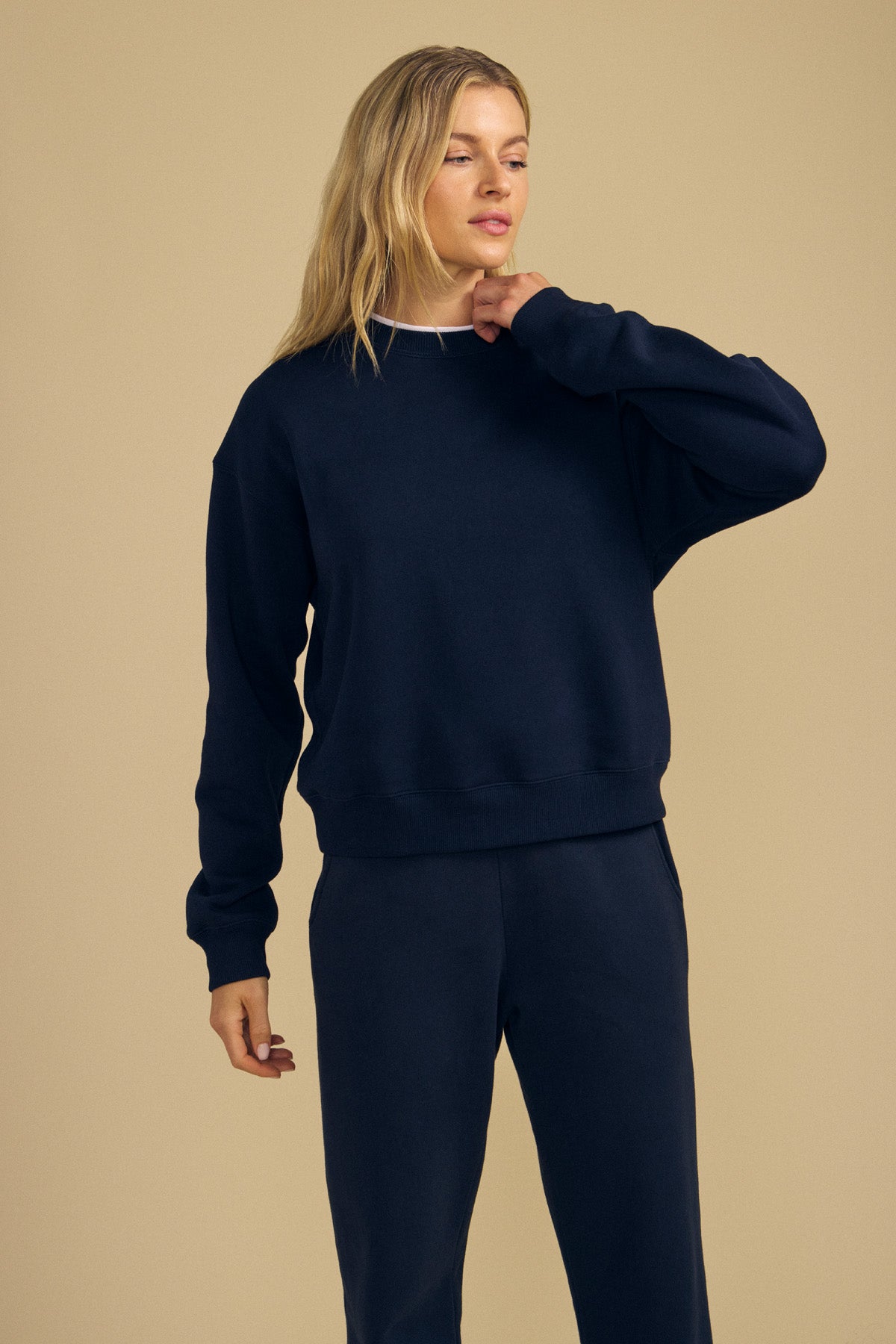 A person with long blonde hair wearing a navy blue YOKO SWEATSHIRT from Velvet by Graham & Spencer and matching soft cotton fleece pants stands against a beige background, showcasing a relaxed silhouette.-42186544906433