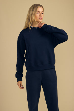 A person with long blonde hair wearing a navy blue YOKO SWEATSHIRT from Velvet by Graham & Spencer and matching soft cotton fleece pants stands against a beige background, showcasing a relaxed silhouette.