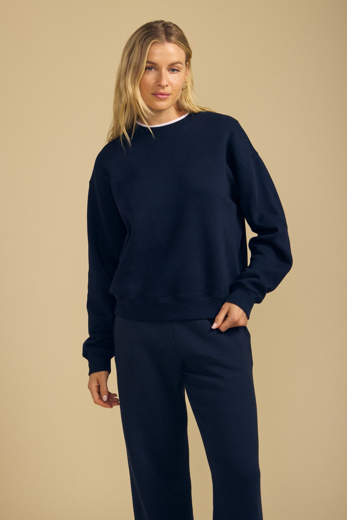 Person standing against a beige background, wearing a navy blue YOKO SWEATSHIRT by Velvet by Graham & Spencer and matching sweatpants crafted from soft cotton fleece, with one hand in a pocket, showcasing a relaxed silhouette.