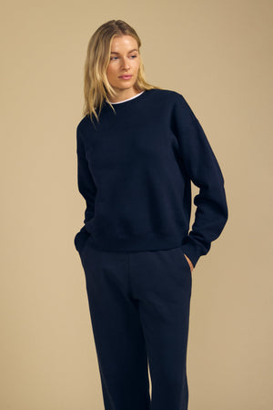 A woman stands with her hands in her pockets against a plain beige background, wearing the YOKO SWEATSHIRT and matching pants from Velvet by Graham & Spencer in dark blue cotton fleece, showcasing a relaxed silhouette.