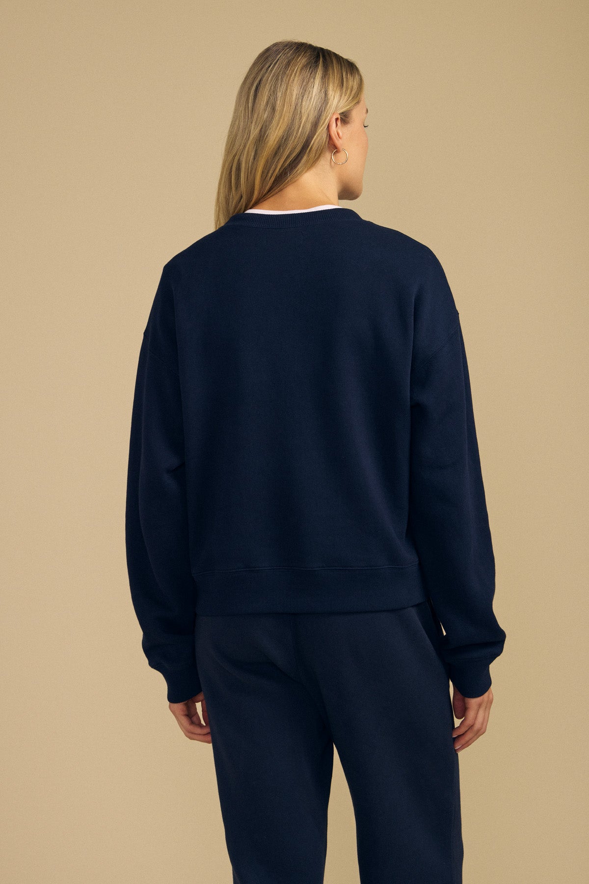 A woman with blonde hair is seen from behind, standing against a beige background and wearing the Velvet by Graham & Spencer YOKO SWEATSHIRT in navy blue paired with matching pants. The outfit is crafted from soft cotton fleece and features a relaxed silhouette.