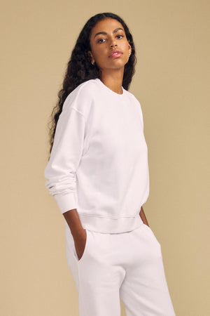 A person with long curly hair wears a white YOKO SWEATSHIRT by Velvet by Graham & Spencer, along with matching sweatpants crafted from soft cotton fleece, standing against a beige background with hands in their pockets, showcasing a relaxed silhouette.