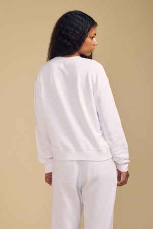 Person with long, curly hair wearing a white YOKO SWEATSHIRT by Velvet by Graham & Spencer and matching pants, featuring a relaxed silhouette in soft cotton fleece, standing and facing away from the camera against a beige background.