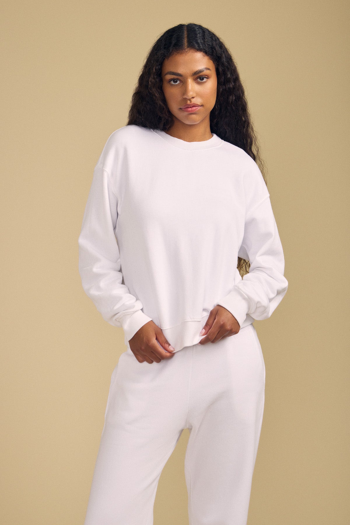 A woman with long curly hair wears the plain white YOKO SWEATSHIRT and matching pants from Velvet by Graham & Spencer, standing against a beige background and gazing at the camera with a neutral expression. The cotton fleece fabric and relaxed silhouette of the sweatshirt lend an effortlessly comfortable feel to her look.